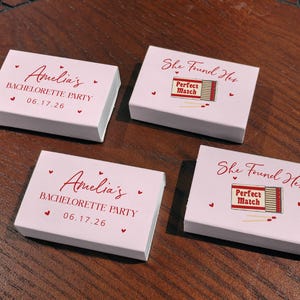 May include: Four white matchbox sets with red text and heart accents. Two boxes read "Amelia's Bachelorette Party 06.17.26." The other two say "She Found Her Perfect Match."