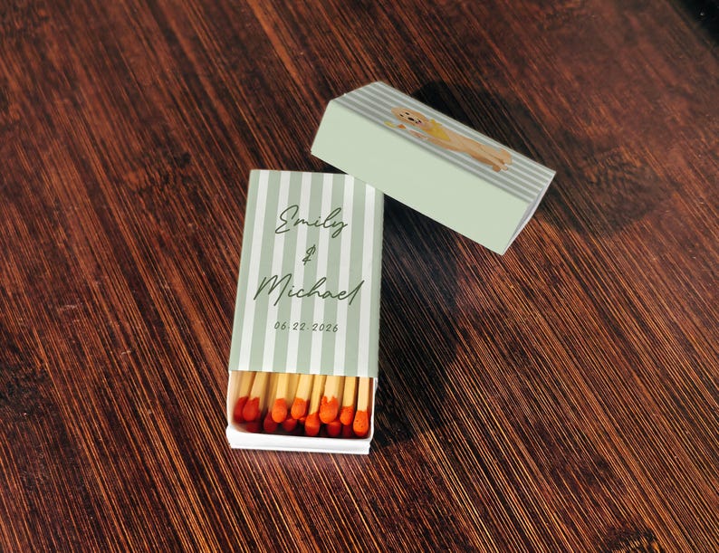 May include: Matchboxes with a light green and white striped design. One box is open, revealing red-tipped matches. The text "Emily & Michael" and the date "06.22.2026" are printed on the front. A second box features a dog illustration.