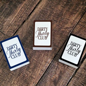 May include: Three decks of cards with the text "DIRTY Thirty CLUB" in a script font. The card decks have white faces with the text in black. The edges of the decks are navy blue, brown, and black. The cards are on a wooden surface.