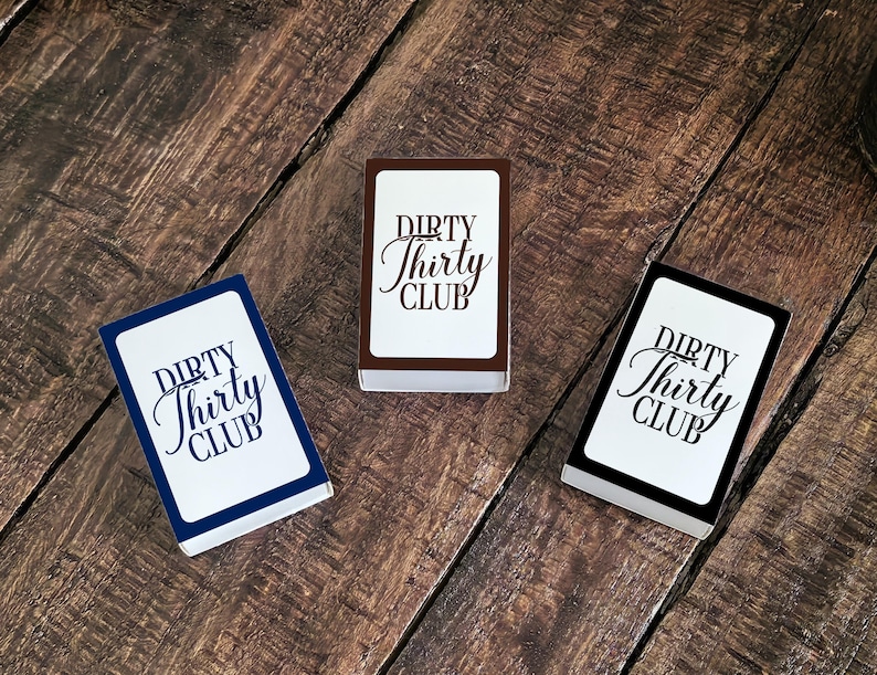 May include: Three decks of cards with the text "DIRTY Thirty CLUB" in a script font. The card decks have white faces with the text in black. The edges of the decks are navy blue, brown, and black. The cards are on a wooden surface.