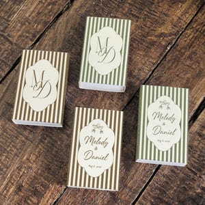 Peut inclure: Quatre bo&icirc;tes d'allumettes rectangulaires &agrave; rayures verticales vert olive ou marron. Deux bo&icirc;tes pr&eacute;sentent le monogramme MD sur une &eacute;tiquette festonn&eacute;e blanche. Deux bo&icirc;tes portent le texte Melody & Daniel avec une date et un motif.