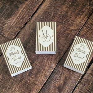 Peut inclure: Trois bo&icirc;tes d'allumettes rectangulaires avec un motif &agrave; rayures verticales blanches et dor&eacute;es. Deux bo&icirc;tes pr&eacute;sentent une &eacute;tiquette blanche avec le texte Melody & Daniel et Aug 6, 2026. La bo&icirc;te centrale porte le monogramme MD en lettres dor&eacute;es.