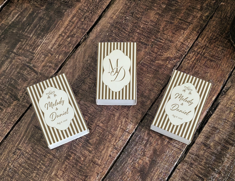Peut inclure: Trois bo&icirc;tes d'allumettes rectangulaires avec un motif &agrave; rayures verticales blanches et dor&eacute;es. Deux bo&icirc;tes pr&eacute;sentent une &eacute;tiquette blanche avec le texte Melody & Daniel et Aug 6, 2026. La bo&icirc;te centrale porte le monogramme MD en lettres dor&eacute;es.