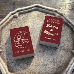 May include: Two rectangular matchboxes with a deep red finish and cream-colored text and graphics. One box features two hands clinking wine glasses with the text 06.18.2026, D+E, and LOS ANGEL,CA. The other box reads I STOLE THIS FROM Diana & Ethan's WEDDING.
