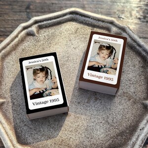 May include: Two rectangular boxes with a photograph of a young child. The boxes have the text "Jessica's 30th" and "Vintage 1995". One box is black, and the other is brown. The boxes are on a gray, textured tray.