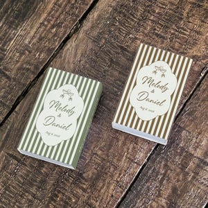 Peut inclure: Deux bo&icirc;tes d'allumettes rectangulaires &agrave; rayures verticales vert olive et marron. Chaque bo&icirc;te pr&eacute;sente une &eacute;tiquette festonn&eacute;e blanche avec le texte Melody & Daniel et Aug 6, 2018, accompagn&eacute; d'un petit graphique de deux fl&ucirc;tes &agrave; champagne.