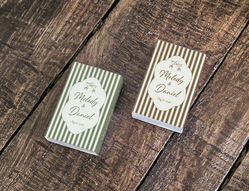 Peut inclure: Deux bo&icirc;tes d'allumettes rectangulaires &agrave; rayures verticales vert olive et marron. Chaque bo&icirc;te pr&eacute;sente une &eacute;tiquette festonn&eacute;e blanche avec le texte Melody & Daniel et Aug 6, 2018, accompagn&eacute; d'un petit graphique de deux fl&ucirc;tes &agrave; champagne.