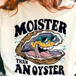 May include: Cream-colored sweatshirt featuring a cartoon oyster wearing teal sunglasses and a wide grin. The text "MOISTER THAN AN OYSTER" is arched above and below the oyster illustration in black lettering. The design includes blue floral accents.