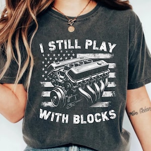May include: Dark gray t-shirt featuring a graphic of an engine and the American flag. The text "I STILL PLAY WITH BLOCKS" is printed in white. The shirt is a vintage style.