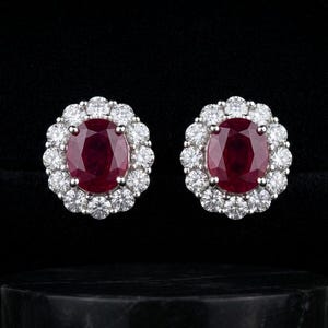 May include: A pair of stud earrings featuring oval-cut, deep red gemstones surrounded by a halo of smaller, sparkling white stones. The earrings are set in a silver or white metal, displayed against a black background.
