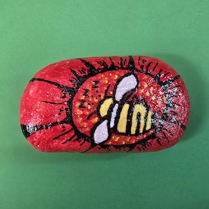 May include: Hand-painted rock featuring a sleeping bee design. The rock is red with a black outline and a yellow and orange center. The bee is yellow and black with white wings, and the word "zzz" is written on the rock.
