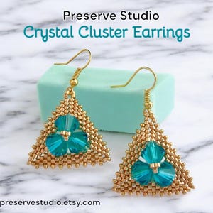 May include: Gold triangle earrings with a cluster of teal blue crystals in the centre. The earrings are on a white marble surface with a teal green box behind them. The text "Preserve Studio Crystal Cluster Earrings" is above the earrings.