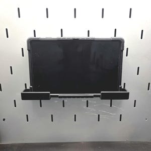 May include: A black tablet in a protective case is mounted on a black pegboard shelf. The shelf has two side supports and a central bar for stability. The pegboard has evenly spaced vertical slots.