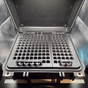May include: A black hard-shell protective case with an open lid, containing two removable black plastic trays with numerous circular cutouts for storage. The trays feature integrated handles for easy removal from the case interior.