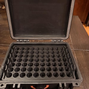 May include: A black hard-shell protective case with an open lid, featuring a custom interior insert with a grid of sixty circular storage slots. The case includes a front-facing circular pressure release valve with orange accents and a sturdy handle.
