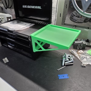 May include: Black US GENERAL tool chest with a green side tray. The tool chest has drawers and an open top. The text "US GENERAL" is printed in white on the top. A collection of tools and hardware are on the surface.