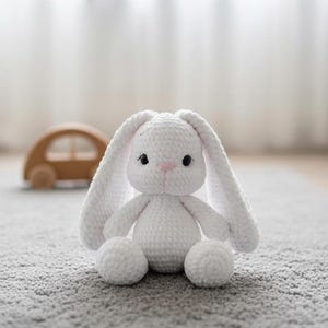 May include: A handmade white crochet bunny with long floppy ears, black eyes, and a pink nose. The plush toy is sitting on a gray carpet. A wooden toy car is in the background.