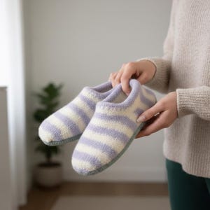 Può includere: Un paio di pantofole a righe bianche e lavanda. Le pantofole hanno un aspetto morbido e strutturato e una suola azzurra. Le pantofole sono tenute nelle mani di una persona.