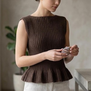 Puede incluir: Top sin mangas de punto marrón con peplum. El top tiene una textura acanalada y un cuello barco. Se sostiene una pequeña bola decorativa multicolor. El top se combina con pantalones de color claro.