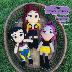 Puede incluir: Tres muñecas de ganchillo hechas a mano con rostros detallados y atuendos coloridos, dispuestas en una cesta de mimbre sobre hierba verde. El texto "KPOP DEMON HUNTERS" y "PDF PATTERN" es visible.