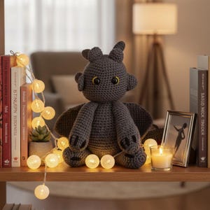 May include: A handmade, dark gray crocheted dragon plush toy with yellow eyes, wings, and small ears. The dragon is sitting on a wooden shelf with a string of glowing globe lights, books, and a framed picture.