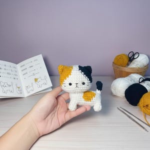 May include: A handmade crochet cat in white, yellow, and navy blue. The cat is held in a hand, with a book of cat diagrams and yarn in the background. The cat has a yellow and navy blue head, yellow spots, and a black tail.