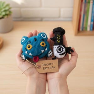 May include: Two handmade crochet toys: a blue monster with yellow eyes and a black and white bird. A tag that says "CROCHET PATTERN" is attached. The toys are held in open hands.