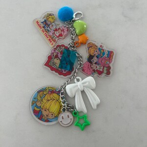 May include: A colorful keychain featuring 80s cartoon characters and charms. The keychain includes a blue pom-pom, green star, orange star, white bow, and a smiley face charm. The text "Made in the 80's" is visible on one of the charms.