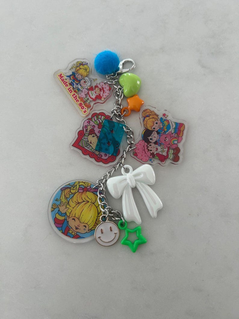 May include: A colorful keychain featuring 80s cartoon characters and charms. The keychain includes a blue pom-pom, green star, orange star, white bow, and a smiley face charm. The text "Made in the 80's" is visible on one of the charms.