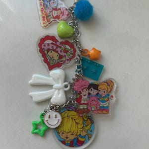 May include: A keychain with multiple charms, including a blue pom-pom, a green heart, and a star-shaped charm. The keychain features several cartoon character charms and the text "Made in the 80's".