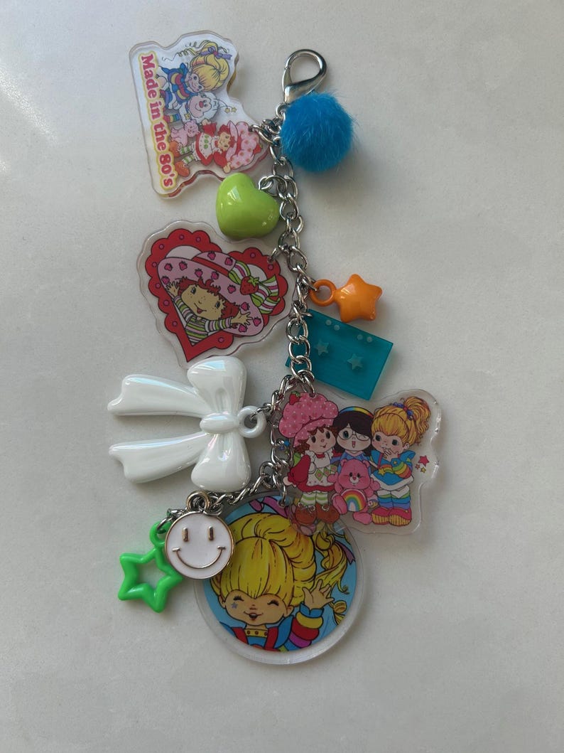 May include: A keychain with multiple charms, including a blue pom-pom, a green heart, and a star-shaped charm. The keychain features several cartoon character charms and the text "Made in the 80's".