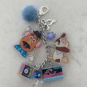 May include: A silver keychain with various charms, including a blue pom-pom, a Mr. Potato Head figure, a dog, a cassette tape, an 8-ball, a star, and a clear gummy bear. The cassette tape features a pink wave design.