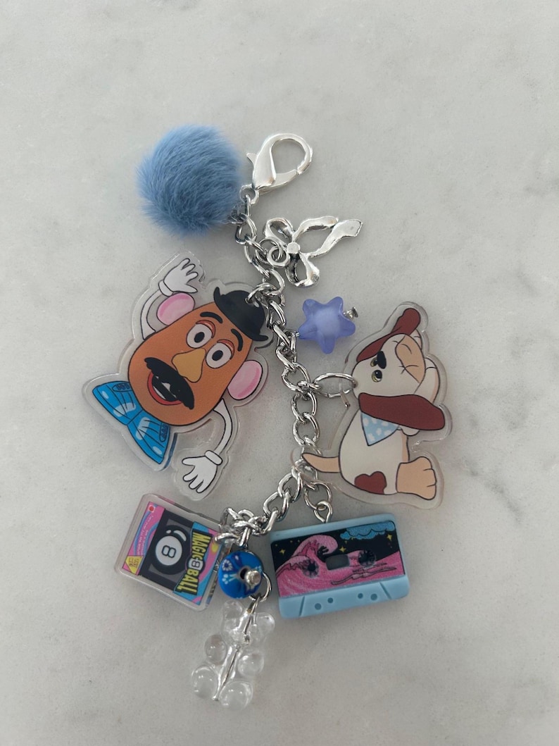 May include: A silver keychain with various charms, including a blue pom-pom, a Mr. Potato Head figure, a dog, a cassette tape, an 8-ball, a star, and a clear gummy bear. The cassette tape features a pink wave design.