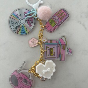May include: A keychain featuring various charms with a retro theme. Charms include a CD with alien graphics, a pink cell phone, a computer, a boombox, and a white heart. The keychain has a gold chain and a pink pom-pom.