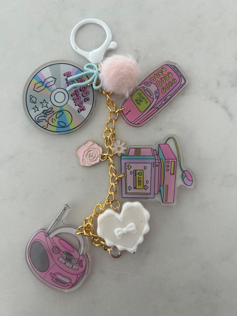 May include: A keychain featuring various charms with a retro theme. Charms include a CD with alien graphics, a pink cell phone, a computer, a boombox, and a white heart. The keychain has a gold chain and a pink pom-pom.