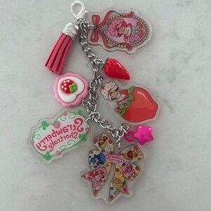 May include: A keychain featuring multiple charms with a Strawberry Shortcake theme. Charms include a pink tassel, a strawberry, a star, and various Strawberry Shortcake character designs. The keychain is attached with a silver chain.