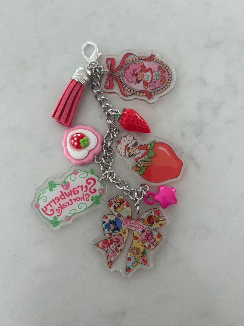 May include: A keychain featuring multiple charms with a Strawberry Shortcake theme. Charms include a pink tassel, a strawberry, a star, and various Strawberry Shortcake character designs. The keychain is attached with a silver chain.