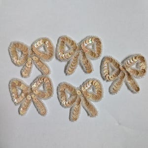 May include: Five decorative bow appliques. Each bow is made of light peach-colored sequins and outlined with small white beads. The bows are arranged on a white background.