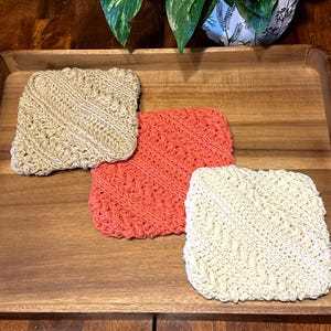 May include: Three square, hand-knitted washcloths in beige, coral, and cream colors. The washcloths are arranged on a wooden tray, showcasing their textured, diagonal stitch pattern. The tray is made of wood.