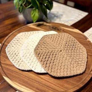 May include: Three crocheted coasters in shades of beige and brown are arranged on a wooden serving tray. The coasters have a hexagonal shape and a textured, open weave design. A potted plant is visible in the background.