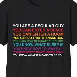 Peut inclure: T-shirt noir avec un motif texte multicolore. Le texte comprend la phrase "YOU ARE A REGULAR GUY" et d'autres affirmations. Le t-shirt est fait d'une matière douce.