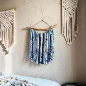 May include: A wall hanging with a natural wood branch and a collection of blue, white, and teal yarn strands. The yarn is arranged vertically, creating a textured, decorative piece. Two macrame wall hangings are also visible.