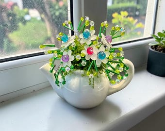 May include: A white ceramic teapot filled with greenery and beaded flowers. The flowers have white petals with colourful centres in pink, blue, and purple. Green beaded stems and leaves extend from the flowers. The teapot sits on a white windowsill.