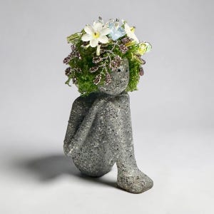 May include: A gray stone sculpture of a seated figure with a floral crown. The figure is in a seated position with its arms wrapped around its knees. The crown features white, blue, and yellow flowers with green foliage.