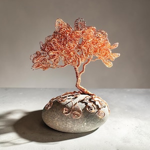 May include: A sculpture of a tree made from twisted copper wire, featuring a dense canopy of coiled wire branches. The trunk of the tree is anchored to a smooth, oval-shaped gray stone, with copper wire roots extending across the surface of the rock.