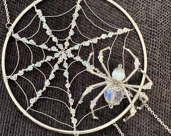 May include: A silver dream catcher with a spider web design, adorned with clear, iridescent beads. A detailed spider with an iridescent bead body and crystal legs is attached to the web. The dream catcher hangs from a delicate chain.