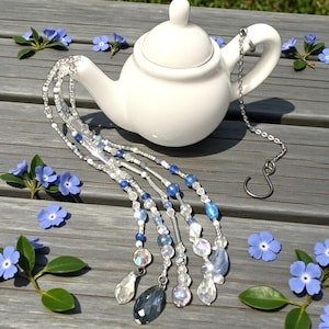 May include: A white ceramic teapot with a lid, adorned with a beaded tea infuser. The infuser features clear, blue, and iridescent beads, with a silver chain. Blue flowers and greenery are scattered around the teapot on a wooden surface.