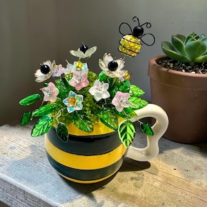 May include: A decorative ceramic mug with a yellow and black striped design, filled with artificial flowers and greenery. The arrangement includes pink, white, and clear flowers, green leaves, and a wire bee. A white handle is attached to the mug.