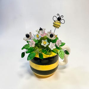 May include: A decorative mug with yellow and black horizontal stripes containing a floral arrangement made of translucent glass flowers, green glass leaves, and a wire-wrapped bee figure with a yellow bead body and black wire wings.