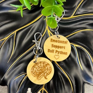 May include: Two wooden keyrings with silver clasps. One keyring has a ball python illustration, the other reads "Emotional Support Ball Python" with a heart symbol. The keyrings are light brown and are on a black surface.